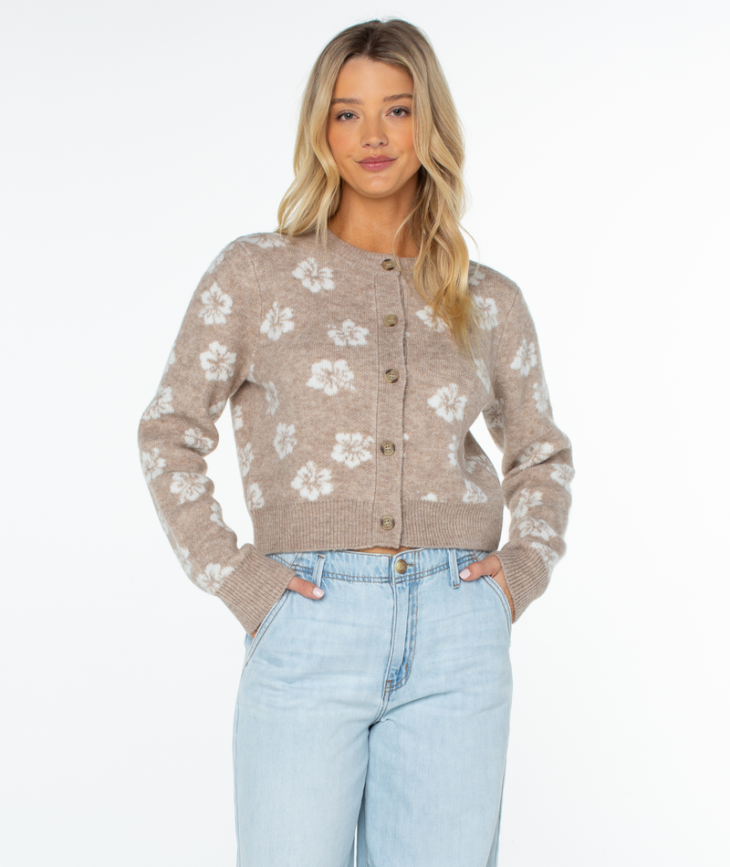 Woman wearing the Meadow Sweater by Roxy in Doeskin Sweet Hibiscus with light blue jeans, hands in pockets, stands against a white background for effortless coastal vibes.