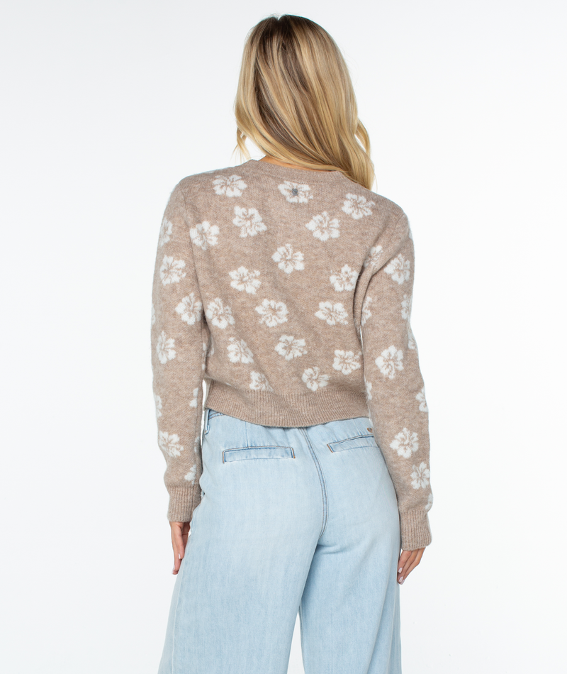 A woman faces away from the camera, wearing the Roxy Meadow Sweater with white floral patterns and light blue high-waisted jeans for an effortlessly coastal look.