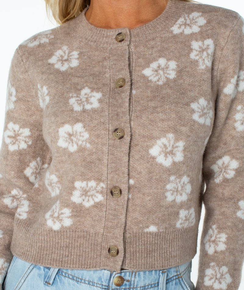 A person wearing the Meadow Sweater by Roxy, a beige button-up cardigan with white floral patterns, styled with light blue jeans for effortless coastal vibes.