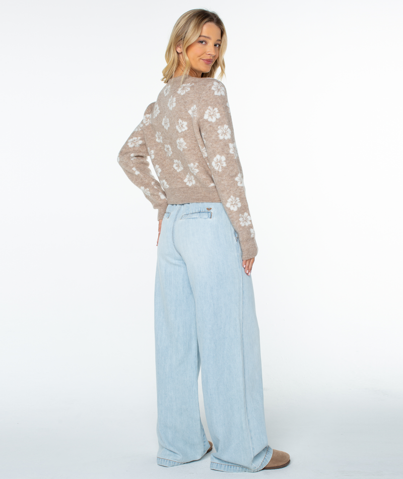 A woman stands smiling and facing backward, wearing the Roxy Meadow Sweater with white floral patterns, paired with light blue wide-leg jeans and beige shoes, creating a relaxed coastal vibe against a white background.
