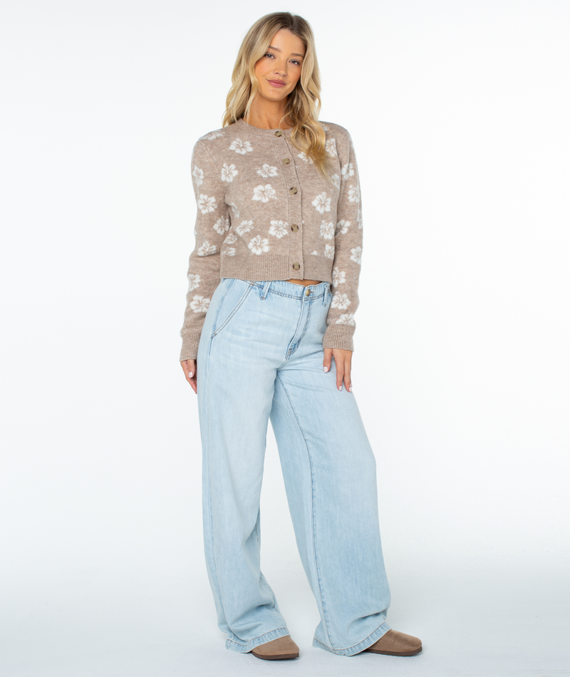 Woman wears the Roxy Meadow Sweater in cozy beige floral knit, paired with light blue wide-leg jeans and beige shoes. She stands against a white background, long blonde hair loose, striking a relaxed pose.