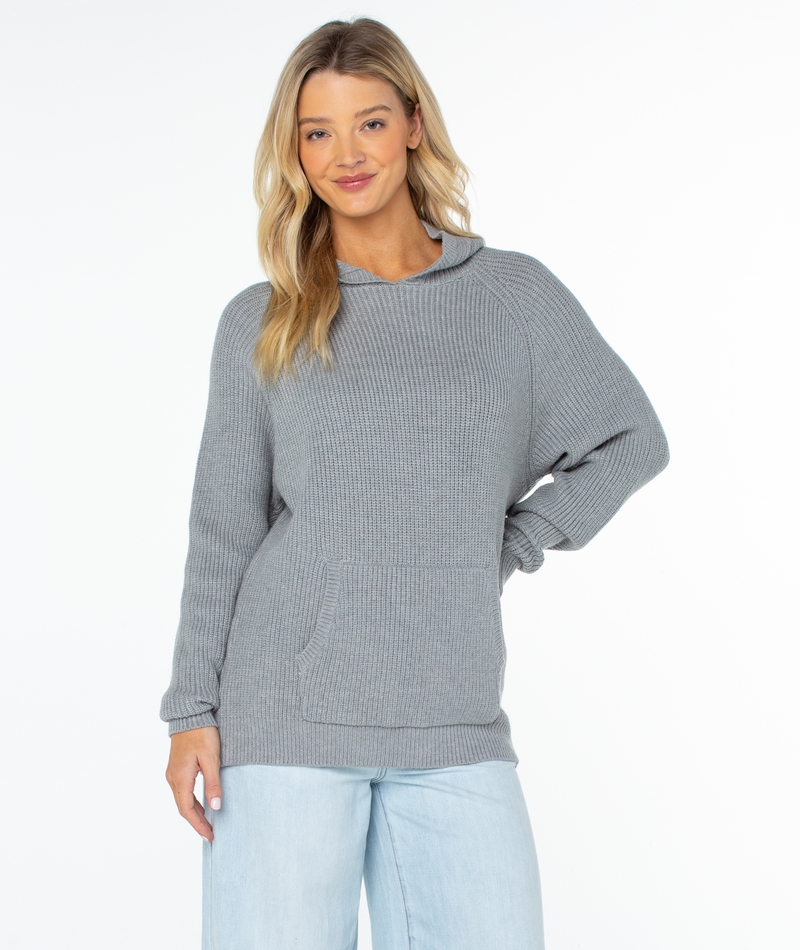 A woman wears a heather grey Roxy Cozy Town Sweater in a knit hoodie style with a front kangaroo pocket and light wash jeans.