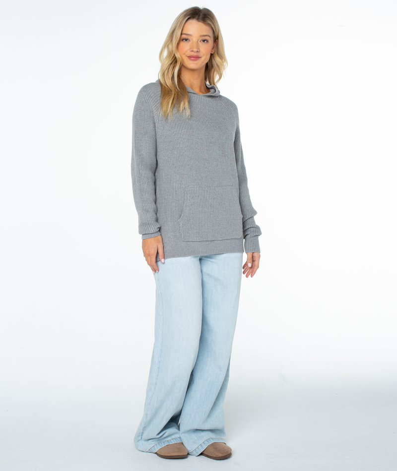 A woman wears a grey, long-sleeve Roxy Cozy Town Sweater with a front pocket and light-wash wide-leg jeans, serving as a relaxed winter staple.