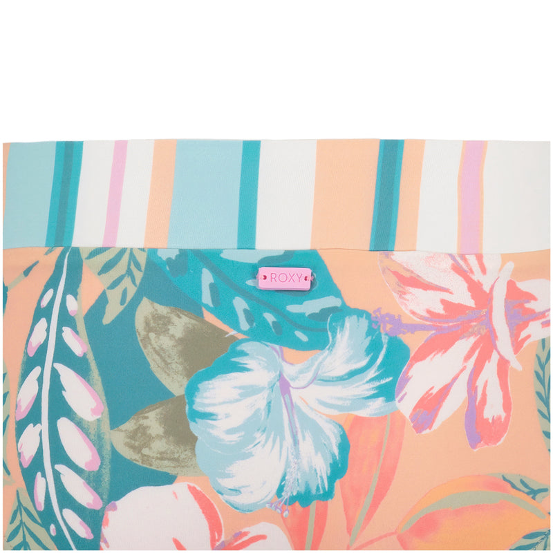 A close-up of the Roxy Girls' Hot Tropics Bikini Set fabric featuring teal, pink, white, and peach floral and leaf patterns with a striped band and a pink Roxy label—ideal for medium coverage swimwear.