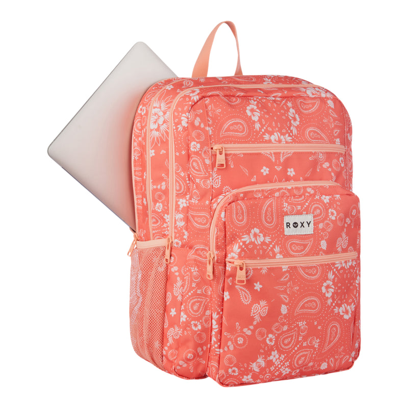 The Roxy Campus Crush Backpack in coral pink is crafted from recycled polyester and displays white paisley patterns. It offers multiple zippered pockets, a mesh side pocket, and a main compartment with a padded laptop sleeve, where a silver laptop can be partially seen.