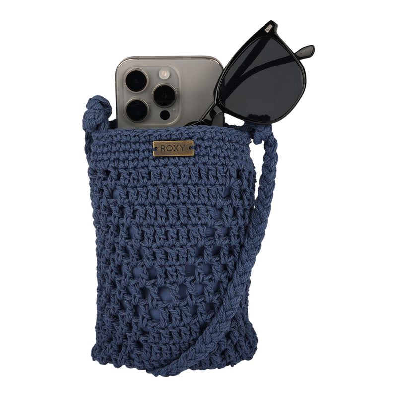 The Roxy Chips And Guac Phone Bag, a blue crocheted crossbody with a long strap and open top, holds a smartphone and black sunglasses—both partially visible—and features the signature ROXY label.