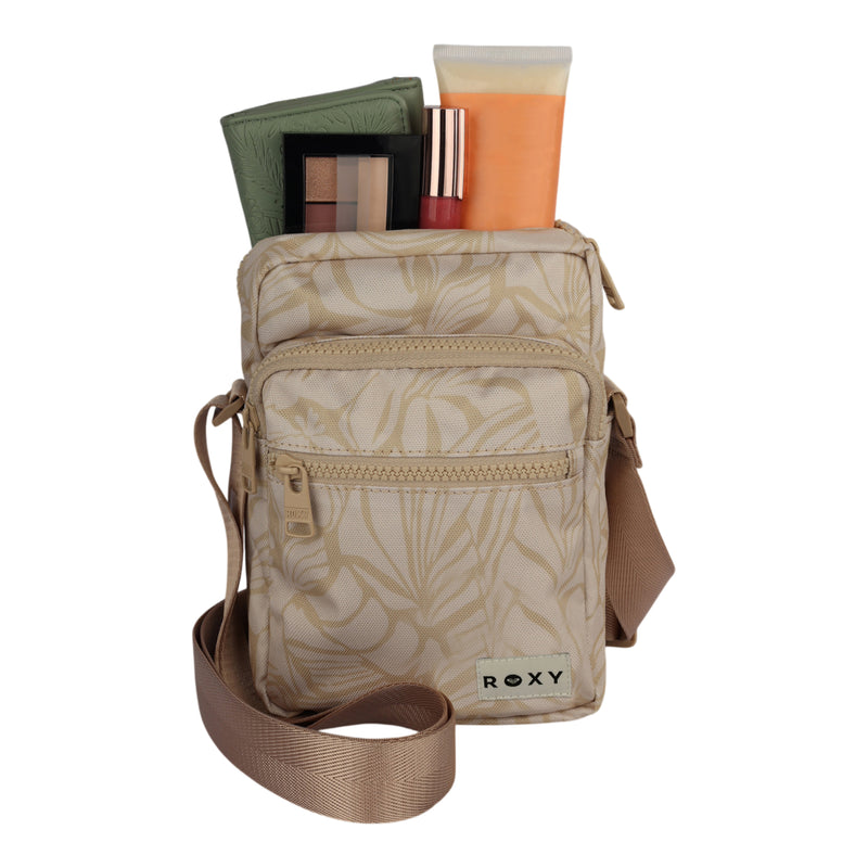 A small beige ROXY Current Mood Crossbody Bag with an adjustable strap and a leaf pattern, holding makeup essentials such as a palette, lipstick, and a cream tube, displayed on a white background.
