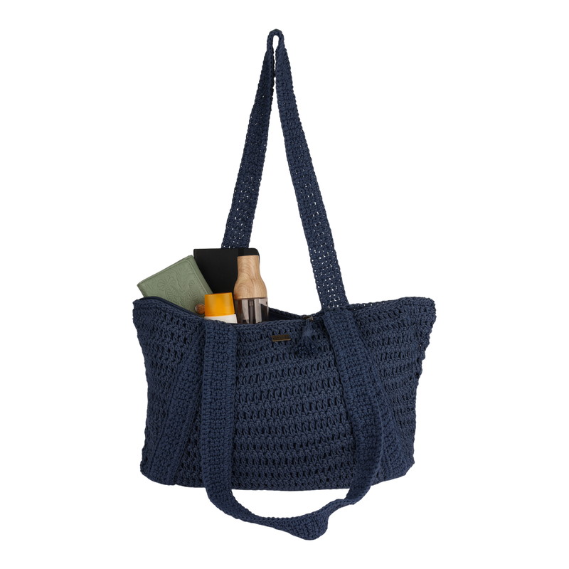 The Chips And Guac Tote Bag by Roxy is a dark blue woven spacious tote with long handles, shown carrying a green book, a black rectangular item, a clear bottle with a wood-colored lid, and a yellow object peeking from the top—making it the perfect travel companion for all your essentials.