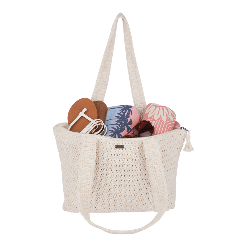 The Chips And Guac Tote Bag by Roxy is a roomy cream crochet beach bag, ideal for travel and stylishly packed with sandals, a pink and blue towel, a patterned pouch, and sunglasses—all set against a white background.