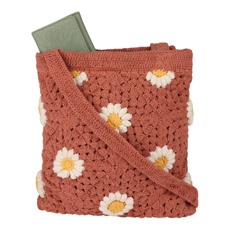 A rust-colored Wandering Beach Crochet Tote Bag by Roxy, featuring white and yellow daisy patterns and carrying a green embossed book partially visible at the top.