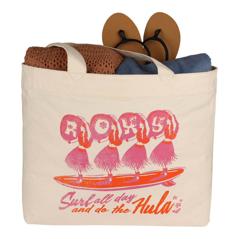 A beige ROXY Surf All Day Tote Bag, featuring eco-friendly materials and pink hula girls with the phrase "Surf all day and do the Hula" printed on it, filled with sandals, a brown towel, and clothing.