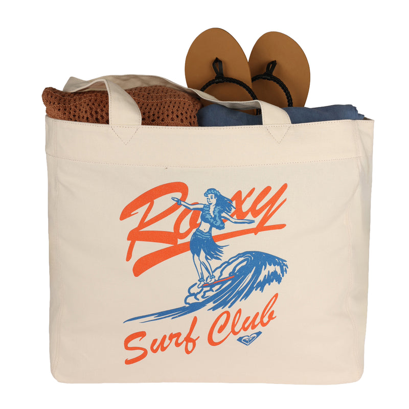 A white ROXY Surf Club Tote Bag featuring a surfer graphic, filled with a brown towel, navy fabric, and tan sandals.
