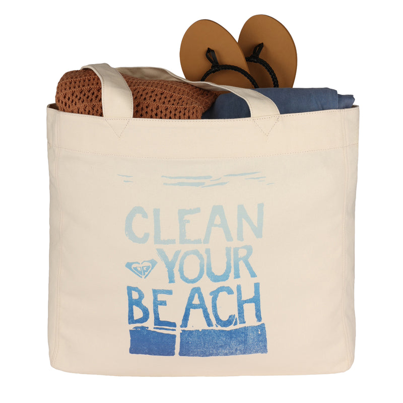 A ROXY Clean Your Beach Tote Bag, featuring "CLEAN YOUR BEACH" text, holds sandals, a brown towel, and a dark blue item.
