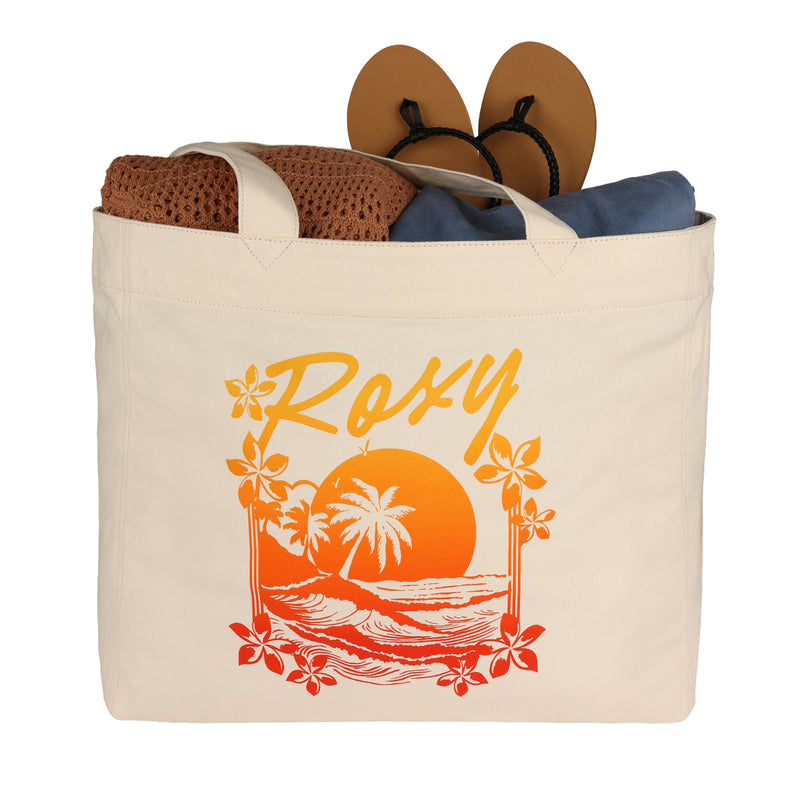 A cream Roxy Sunset Tote Bag by ROXY, decorated with a sunset beach design, shown filled with a brown crochet item, blue fabric, and tan sandals with black straps visible.