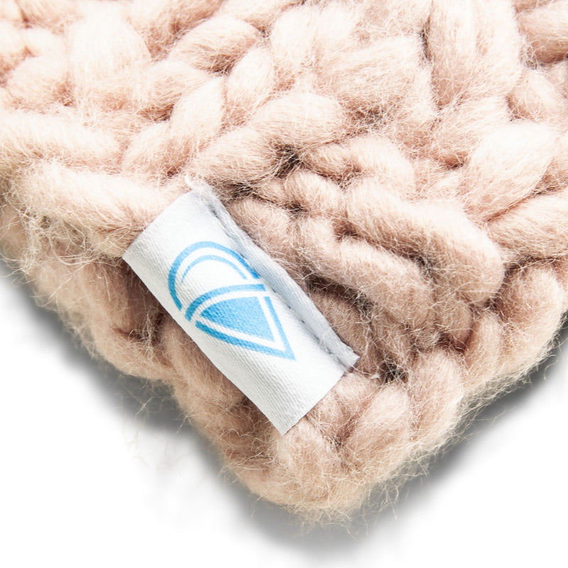 Close-up of the Roxy Winter Collar, a chunky knit neck warmer in pale pink, featuring a white tag with a simple light blue heart-shaped logo. The white background highlights the texture and color of this stylish women’s accessory.