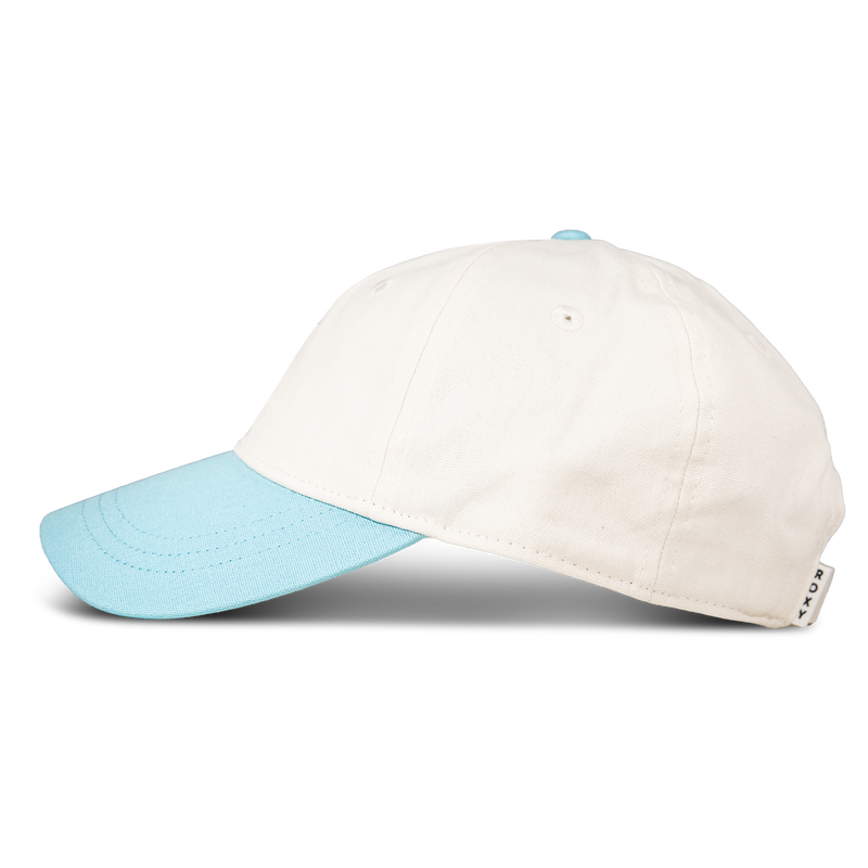 The Roxy Next Level Cap is a women’s baseball hat made from soft cotton twill in white, featuring a light blue curved visor and matching button, shown from the side on a plain white background.