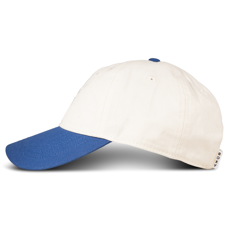 A side view of the Roxy Next Level Cap, a women’s baseball hat featuring a white cotton twill crown and a blue curved visor. A small label with text is visible on the back. The background is plain white.