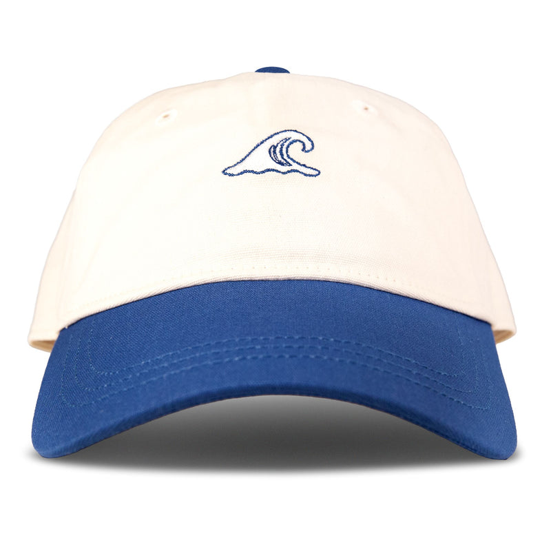 The Roxy Next Level Cap is a women’s baseball hat in Dutch Blue/Wave, featuring a cream-colored cotton twill crown and a blue curved visor. An embroidered dark blue ocean wave icon accents the center front. The cap is displayed isolated against a plain white background.