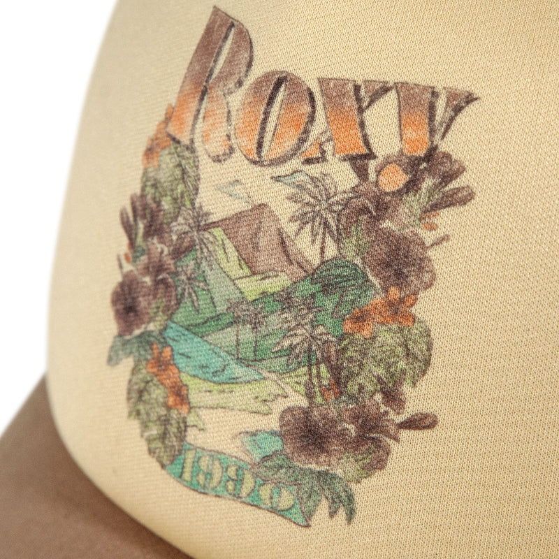 A close-up of the Sandy Top Trucker cap by Roxy in beige, featuring “Roxy” in bold letters above a vibrant tropical graphic with palm trees, flowers, a mountain, and the number 1990. Designed with an adjustable snapback for comfort and sun protection.