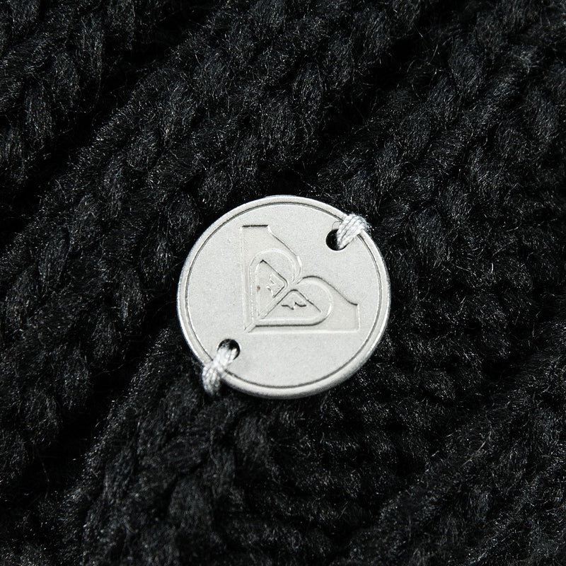 Close-up of a silver circular metal tag stitched onto the thick black knit fabric of a Roxy Blizzard Beanie.