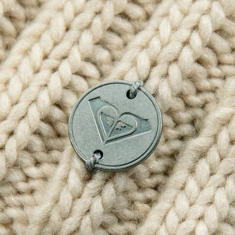 A round, metallic logo tag attached to the chunky knit fabric of a ROXY Blizzard Beanie.