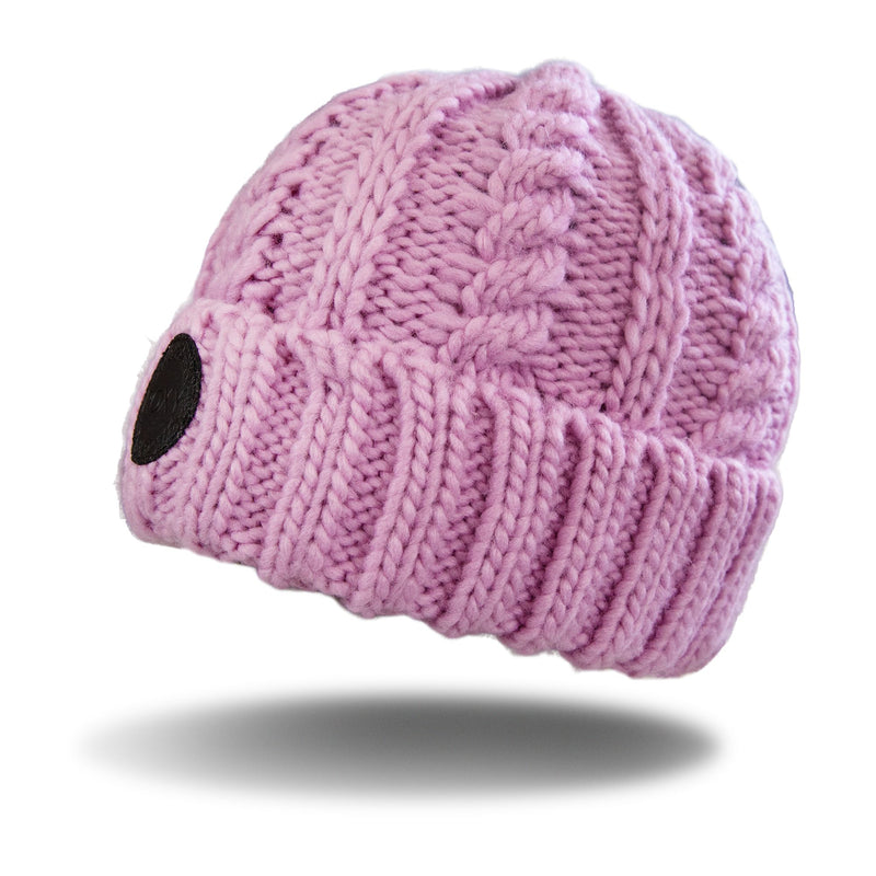 The Tram Beanie by Roxy is a pink, cable knit beanie featuring a thick ribbed cuff and a black circular patch on one side, displayed against a white background with a shadow beneath it.
