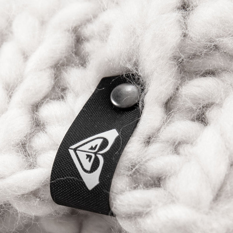A close-up of the Roxy Winter Beanie showcases its thick, white knit design and cozy fleece lining, perfect for women. A black tag, attached with a metal rivet, features Roxy’s signature white heart-shaped abstract logo.