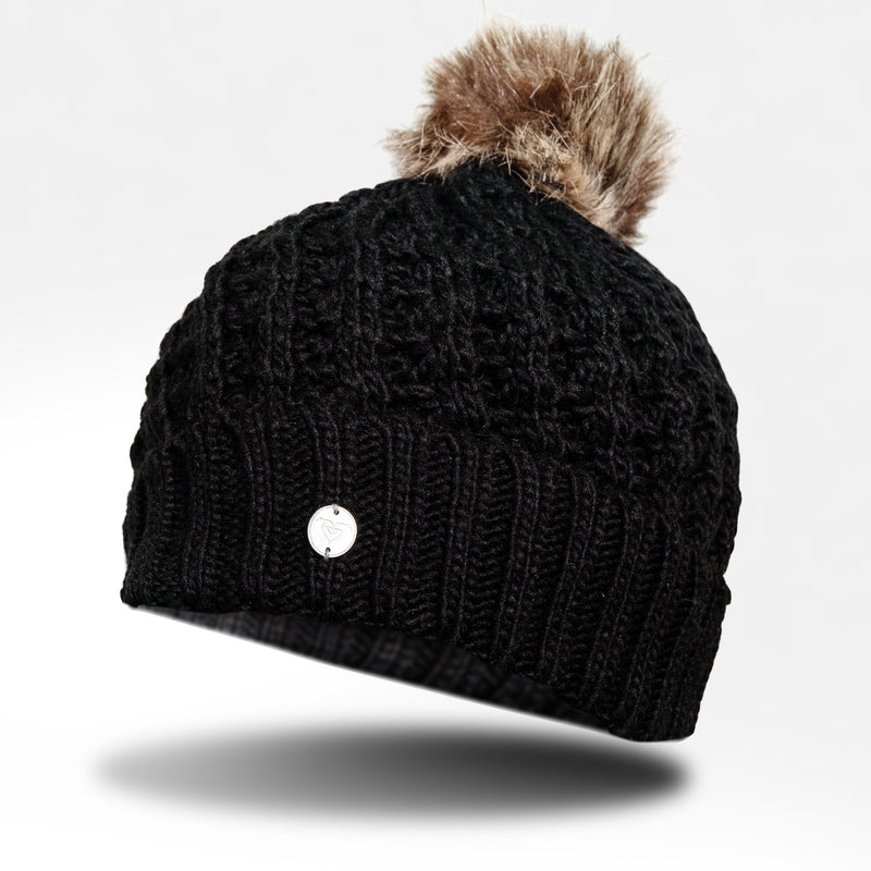 The Roxy Girls Blizzard Girl Beanie by Roxy features a black cable knit design with a brown faux fur pom-pom on top and is finished with a small round silver emblem on the front. The beanie is shown isolated on a white background with a subtle shadow underneath.