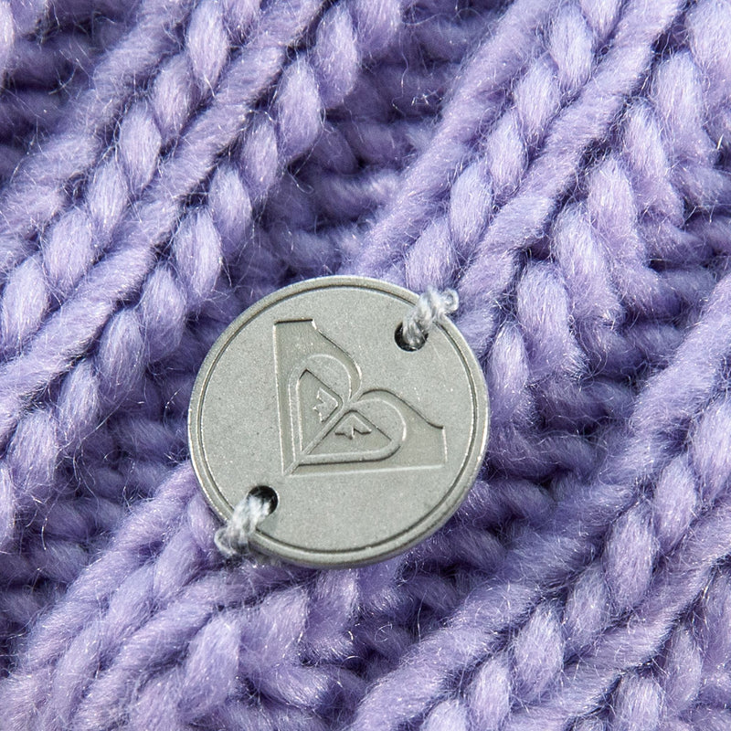 Close-up of a round metal button with a heart-shaped logo, sewn onto the thick, textured, purple cable knit fabric of the Roxy Girls Blizzard Girl Beanie by Roxy, featuring a cozy polar fleece lining.