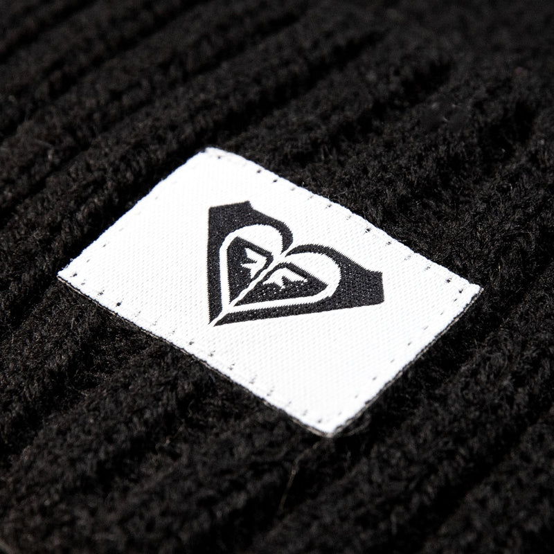 Close-up of a white rectangular label with a black heart-shaped logo stitched onto a black rib knit Roxy Girls So Peaceful Beanie.