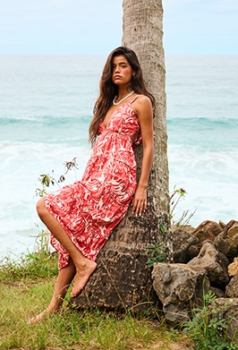 Woman in a pink floral dress standing next to a palm tree by the ocean.