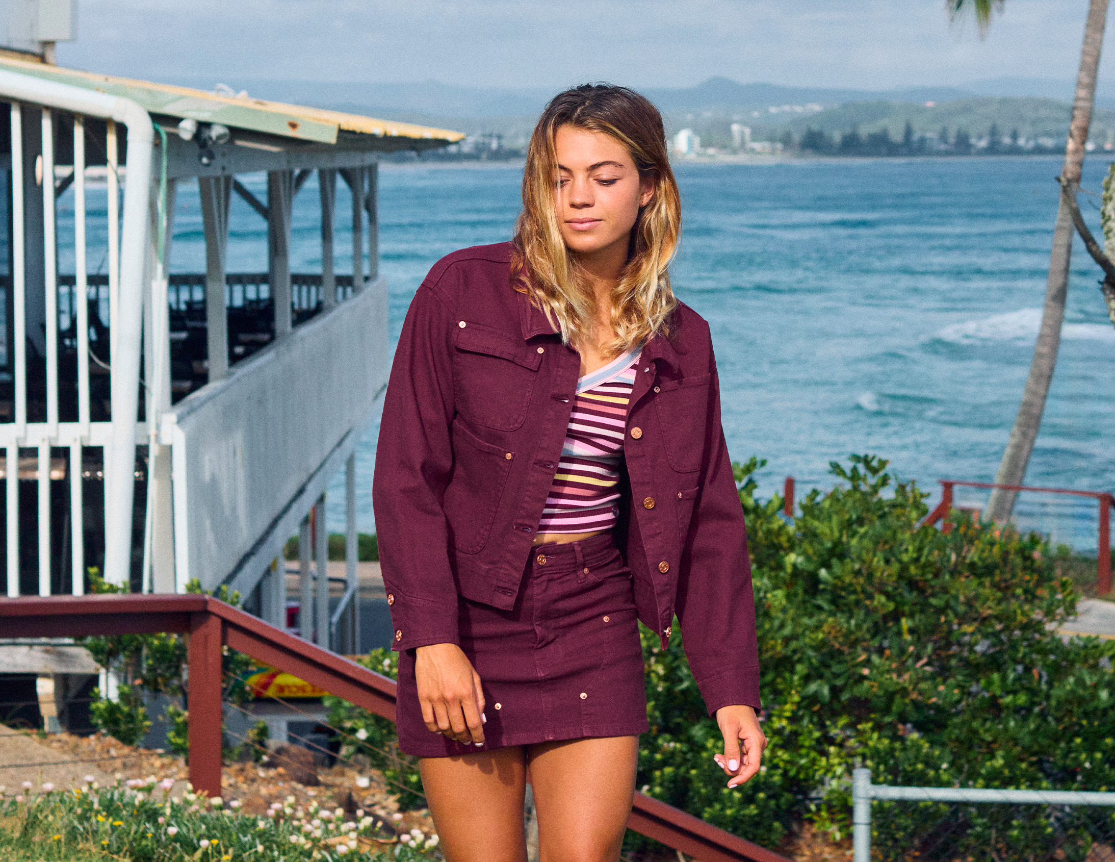 A woman stands outdoors by the ocean wearing a matching burgundy denim jacket and skirt set over a striped top. The background features a coastal pier, greenery, and the blue sea.