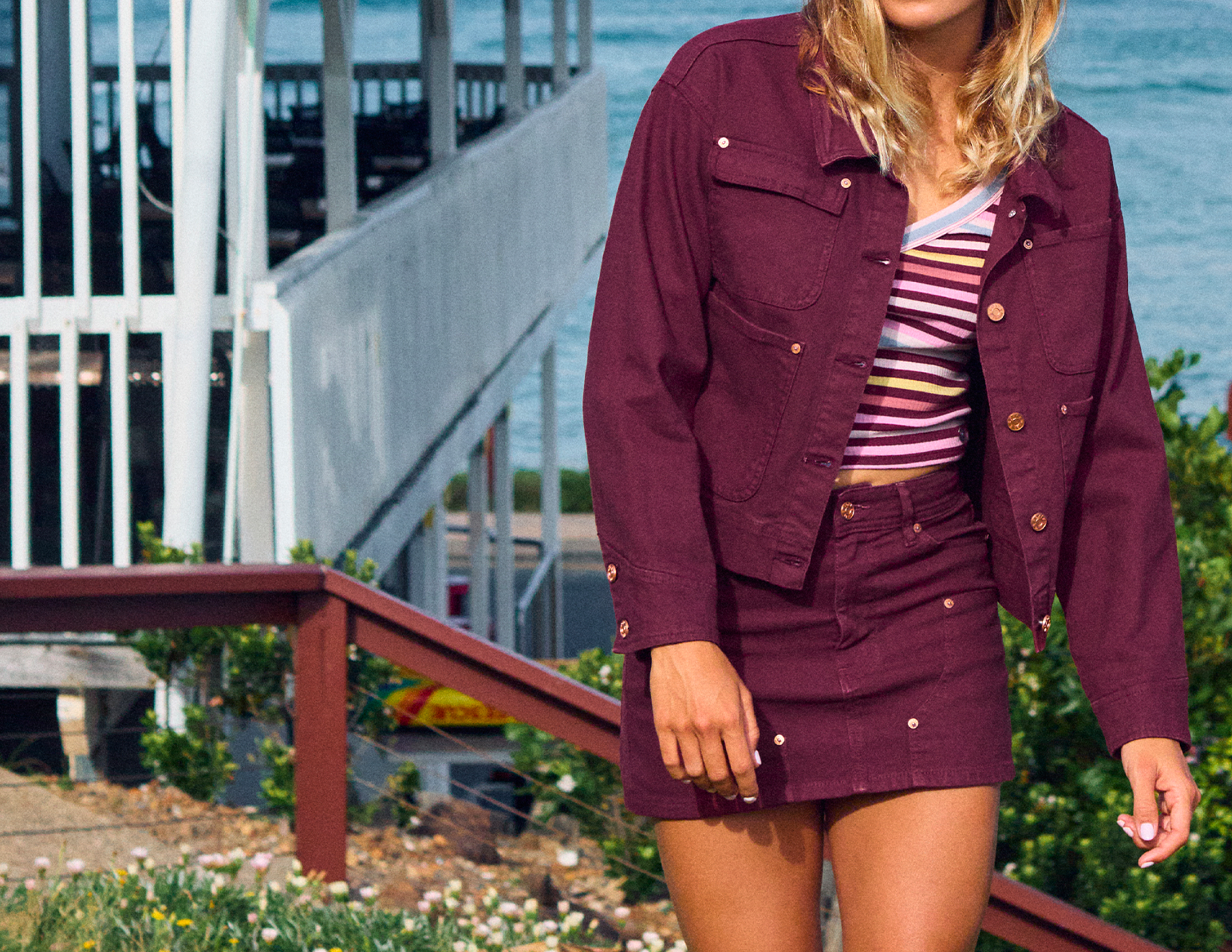 A woman wearing a matching burgundy denim jacket and mini skirt set, styled over a striped top, standing outdoors near a railing and water.