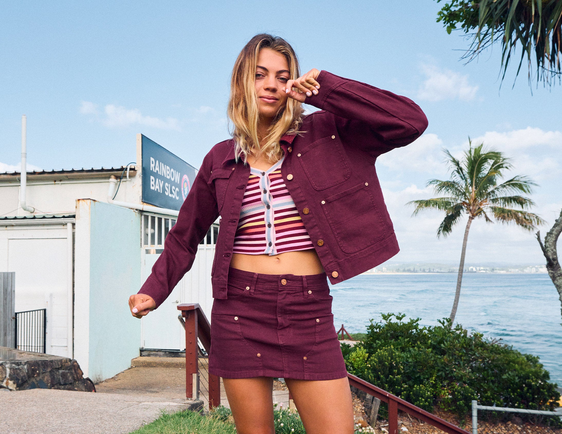 A woman posing outdoors in a matching burgundy denim jacket and skirt set, styled with a striped crop top, with a beach and ocean background.