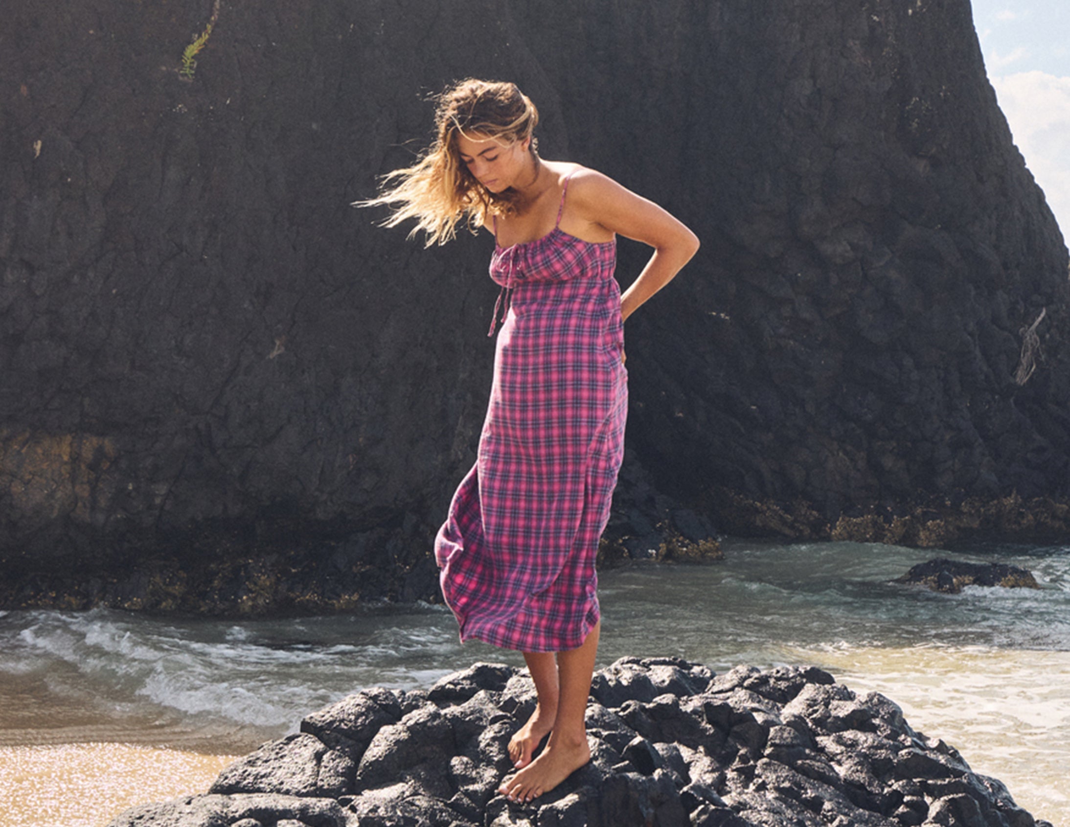 A woman stands barefoot on dark volcanic rocks by the ocean, wearing a pink and purple plaid halter maxi dress with a tie at the chest.