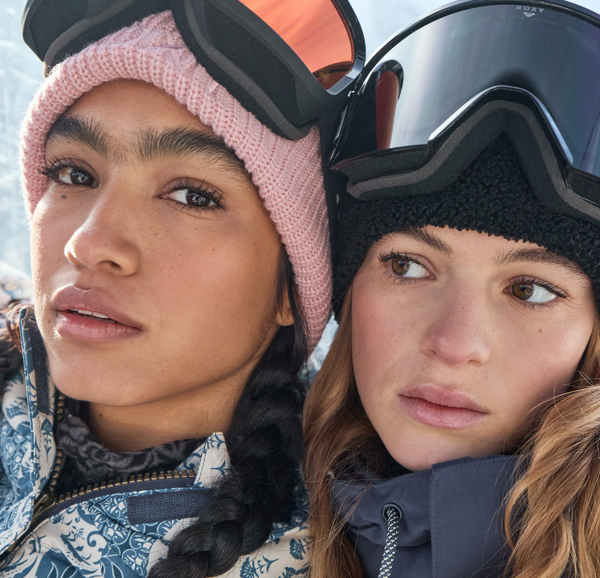 Two women in close-up wearing winter sports gear, including ski goggles and knit beanies, with snow visible in the background.