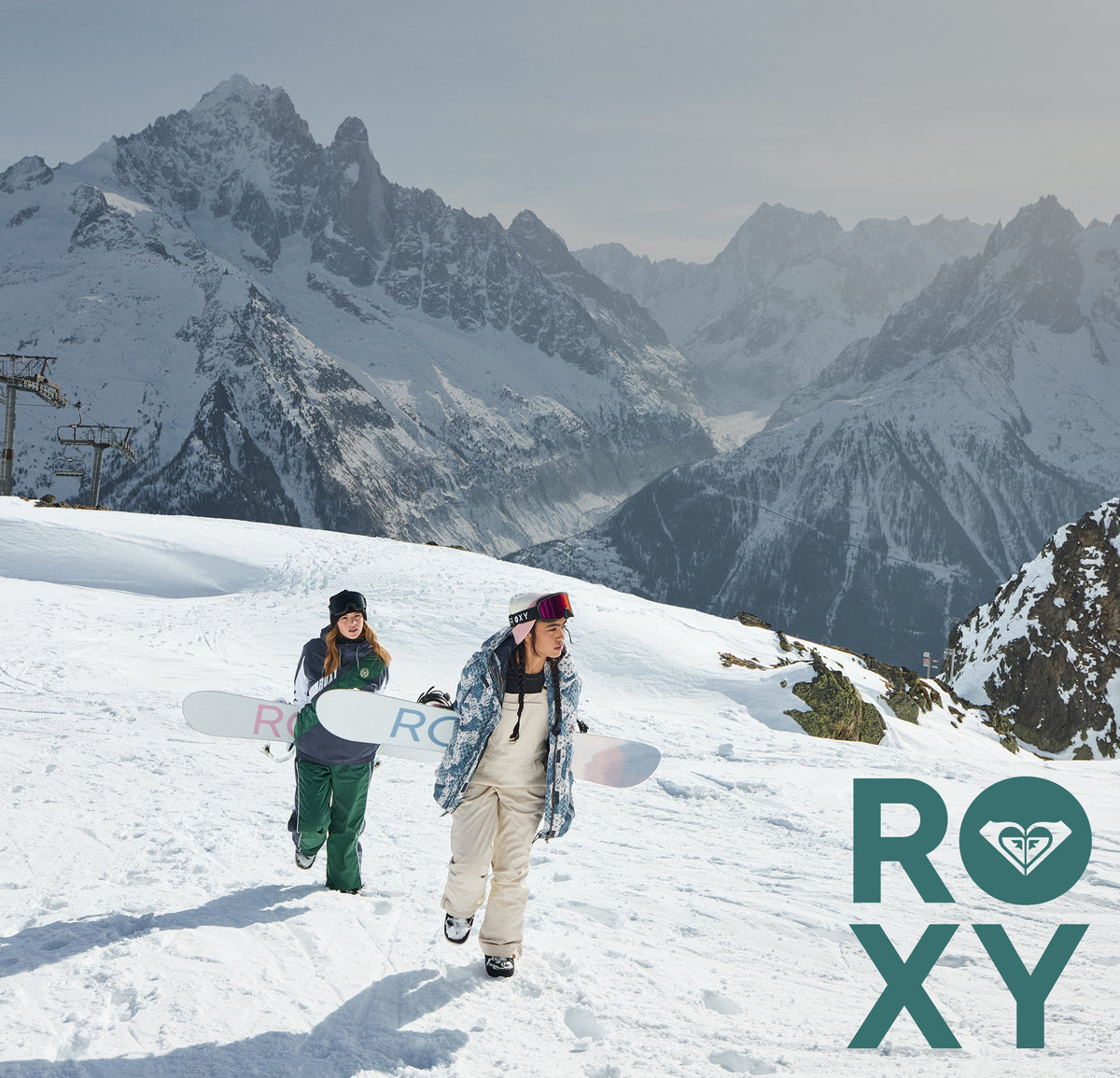 Two snowboarders walking on a snowy mountain slope with dramatic rocky peaks in the background. One person wears a green snowsuit and holds a snowboard, while the other wears a blue plaid jacket over a white top and beige pants.