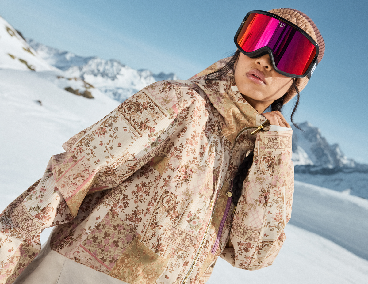 Woman wearing a cream floral snow jacket, knit beanie and pink mirrored goggles in snowy mountains.