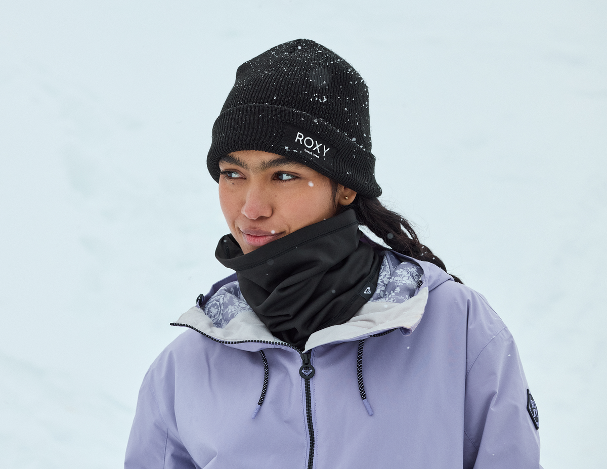 A woman wearing a black Roxy knit beanie with snowflakes, a black neck gaiter, and a light lavender ski jacket, set against a snowy white background.