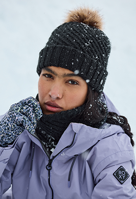 A close-up of a person outdoors in the snow wearing a dark knit pom-pom beanie, a black neck warmer, patterned gloves, and a light purple winter jacket with a visible Brixton logo patch on the sleeve.