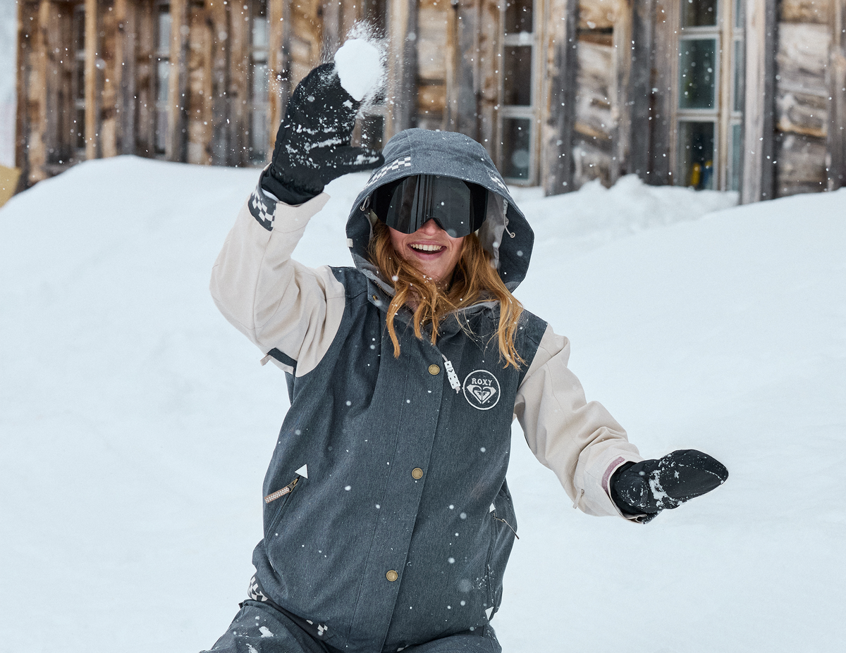 A person snowboarding outdoors in the snow, wearing a dark gray and beige snowboard jacket with black gloves and goggles, with a wooden building in the background.
