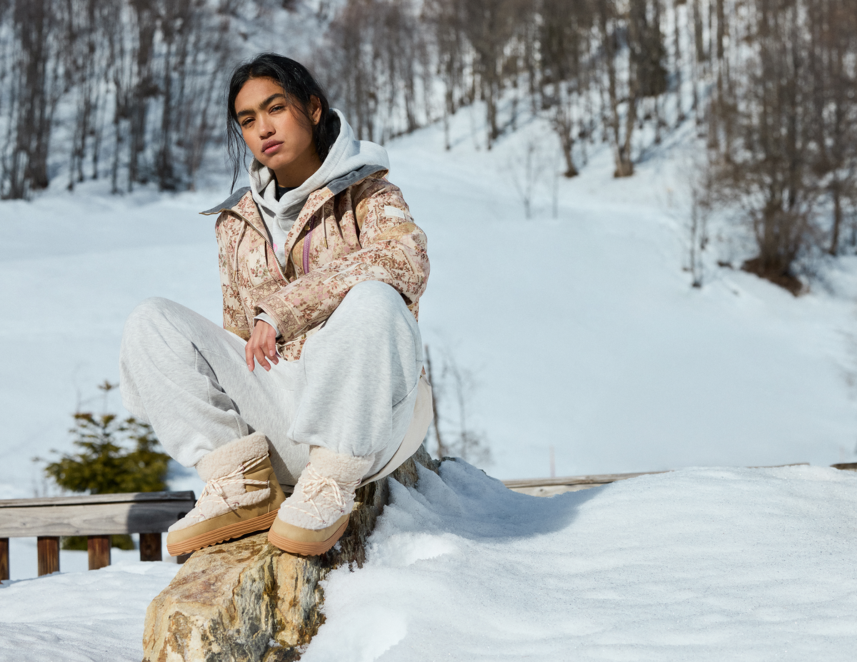 A person sitting outdoors in a snowy landscape wearing a beige and white patterned jacket, light gray sweatpants, and beige boots with white faux-fur trim.
