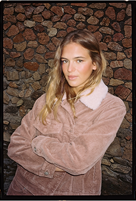 A woman with blonde hair wearing a dusty pink corduroy jacket with a white faux-fur collar, standing in front of a rustic stone wall.