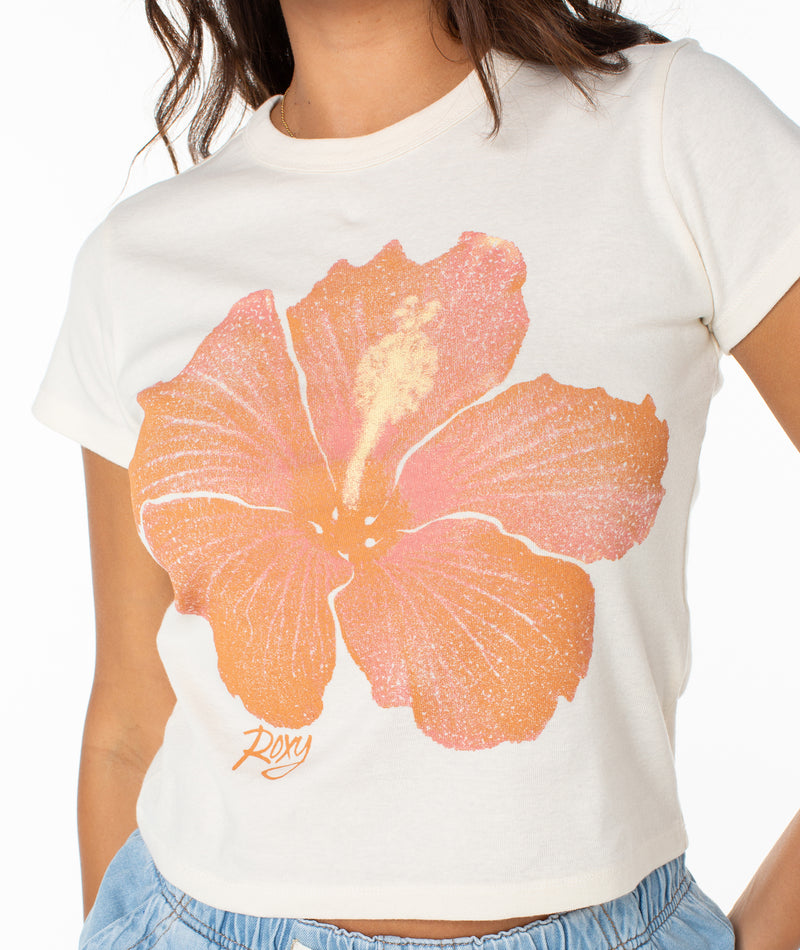 A person wears the Roxy Hibiscus Spray 90S Baby Tee, a cream fitted t-shirt with a bold orange and pink hibiscus flower graphic and "Roxy" below, paired with light blue pants; the person's face is not visible.