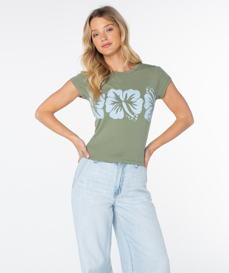 A woman with long blonde hair wears the Roxy Flower Block 90S Baby Tee—a fitted women’s t-shirt in green with bold white hibiscus flowers—paired with light blue jeans, standing against a plain white background for a fresh, retro beachwear-inspired look.