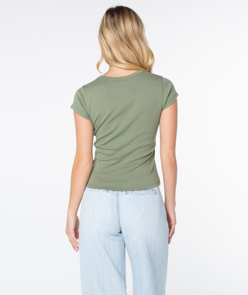 A woman with long blonde hair, standing with her back to the camera against a plain white background, wears the Roxy Flower Block 90S Baby Tee—a fitted, short-sleeve women’s t-shirt in olive green—paired with light blue jeans.