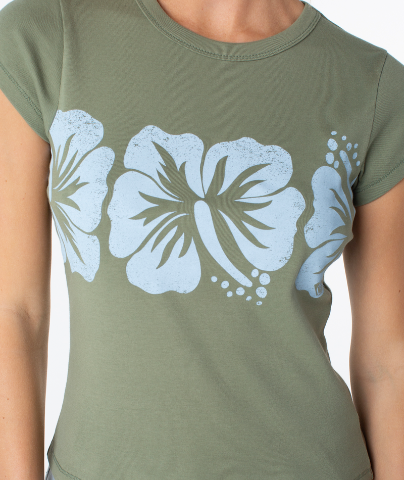 A person wearing the Roxy Flower Block 90S Baby Tee, an olive green fitted women’s t-shirt featuring large, light blue hibiscus flower prints across the chest, evokes a touch of retro beachwear style.
