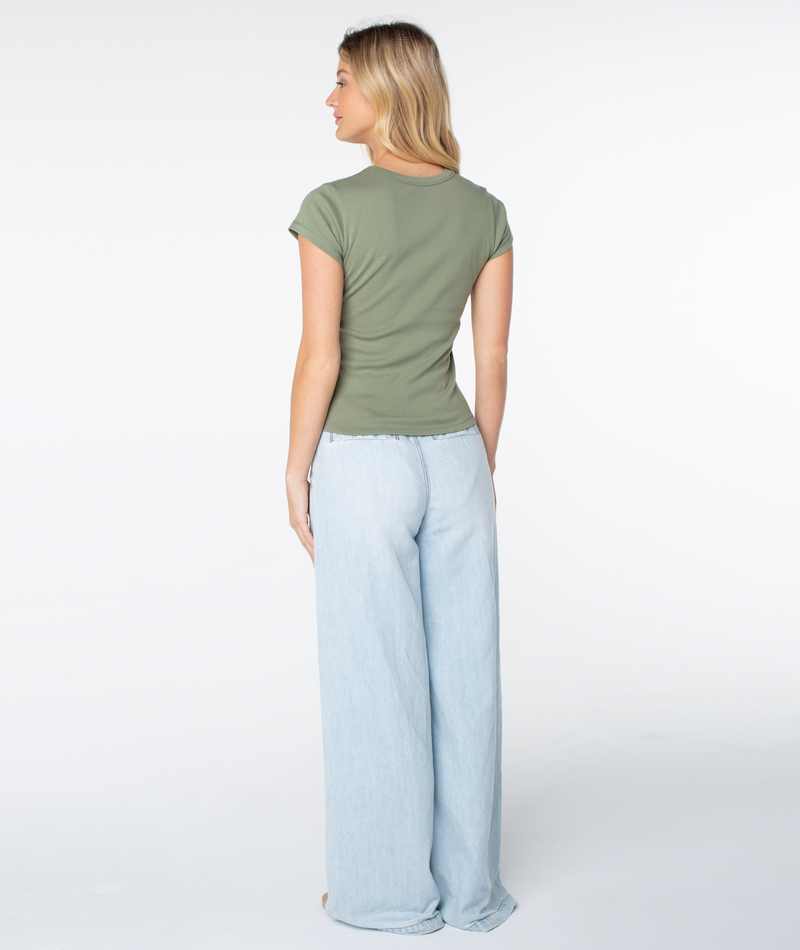 A woman with long blond hair is standing facing away, wearing the Roxy Flower Block 90s Baby Tee in olive green and loose, light-wash wide-leg jeans against a plain white background.