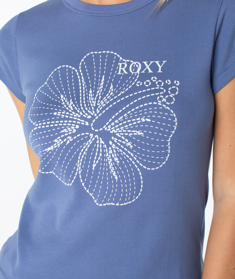 A person wearing the Roxy Stitched Bloom 90S Baby Tee, a retro blue fitted shirt featuring a white stitched hibiscus outline and "ROXY" on the front.