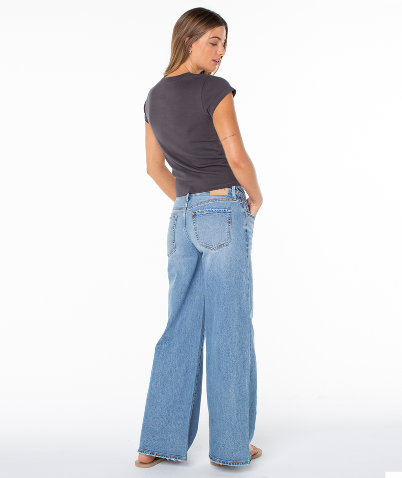 A woman faces away in the Hibiscus View 90S Baby Tee by Roxy and light blue wide-leg jeans, one hand in her pocket, glancing over her shoulder against a white background.